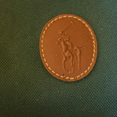 Close-up detail of an embossed leather Polo Ralph Lauren logo patch on green canvas luggage or bag.