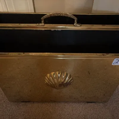 Brass magazine rack featuring a central shell motif and a top carrying handle, showing light tarnish and surface wear.