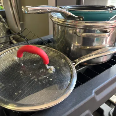 Assorted modern cookware, including a stainless steel pot, non-stick skillets, and a glass pot lid with a red silicone handle.