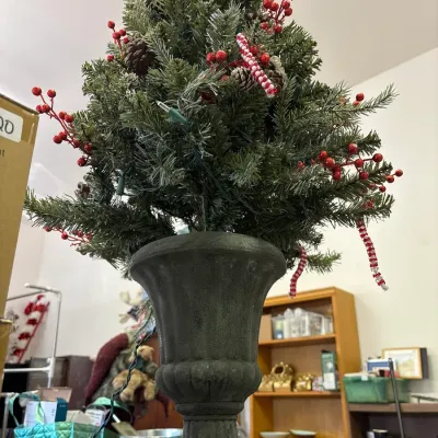 Small pre-lit artificial Christmas tree set inside a dark urn-style planter, decorated with pinecones, red berry sprigs, and candy cane...