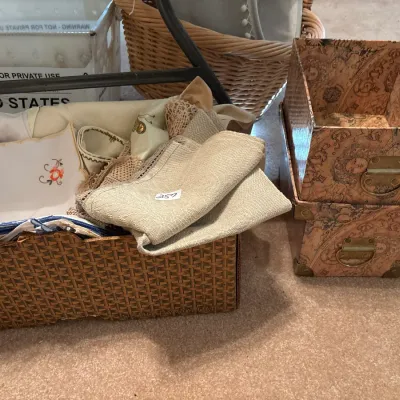 Collection of linens and embroidered handkerchiefs arranged in a woven basket alongside decorative map-patterned storage boxes.