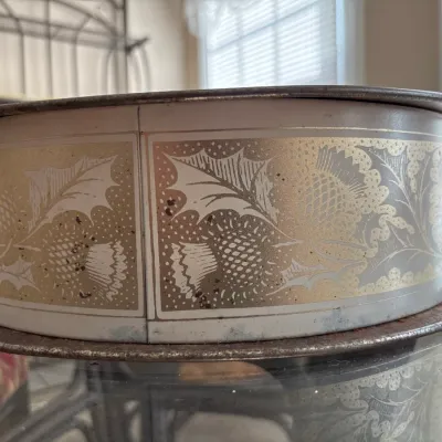 Vintage circular metallic tin container decorated with a gold and silver thistle pattern, resting on a glass tabletop.