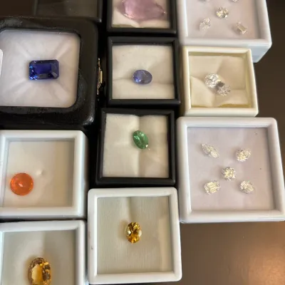 Collection of loose faceted gemstones displayed in individual square presentation cases, including tanzanite, yellow citrine, green...