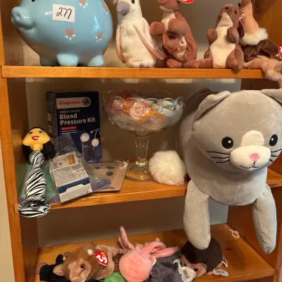 Wooden display shelving unit filled with plush Ty Beanie Babies, a large grey cat plush, a blue piggy bank, a Walgreens blood pressure...