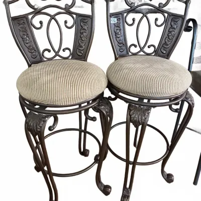 Pair of ornate wrought iron swivel barstools featuring scrollwork backs, leaf-motif details, and padded beige corduroy cushion seats.