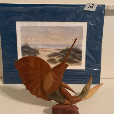 Hand-carved wooden sailfish figurine mounted on a base, displayed alongside a matted coastal landscape art print.