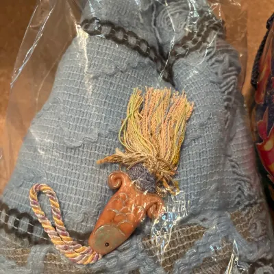 Handcrafted terracotta ceramic fish motif ornament with a multicolored thread tassel, packaged inside a clear plastic sleeve over light...