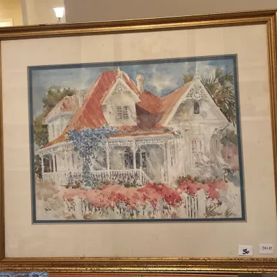Framed watercolor painting depicting a Victorian house with a red roof and wraparound porch, signed by the artist in the lower right corner.