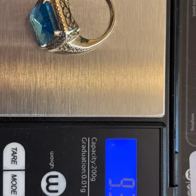 Gold-tone cocktail ring featuring a cushion-cut blue stone with halo accent detail, shown on a digital scale weighing 9.34 grams.