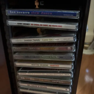 Collection of music CDs in jewel cases stored in a vertical rack, featuring artists like Shania Twain, Kenny Chesney, Celine Dion, and...