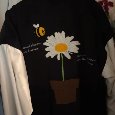Black and white varsity-style jacket featuring chenille patches of a potted daisy and a bumblebee, along with embroidered script text on...