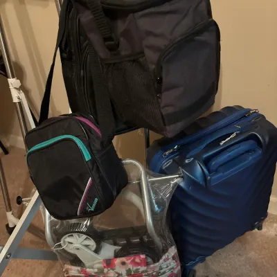 Collection of travel gear including a dark grey cooler backpack, a blue hard-shell rolling suitcase, a small cross-body bag, and a...