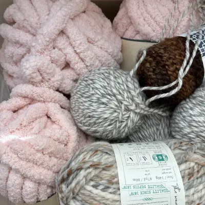 Assorted skeins and balls of knitting yarn, featuring light pink chenille style yarn and Lion Brand super bulky variegated yarn.