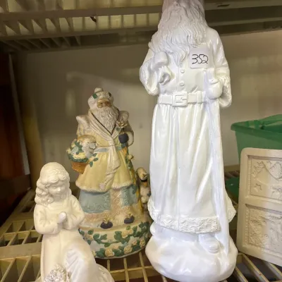 Collection of decorative holiday figurines, including a tall white unpainted Santa Claus statue, a colorful detailed Santa figure, and a...