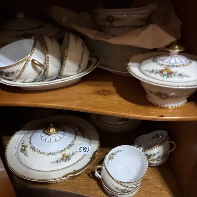 Vintage porcelain dinnerware pieces featuring floral borders and gold gilt accents, arranged on wooden cabinet shelves.