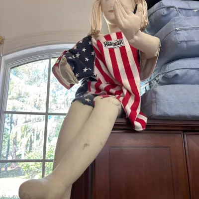 Vintage life-sized soft sculpture cloth doll with yarn pigtails, wearing an American flag theme top, perched atop a wooden cabinet.