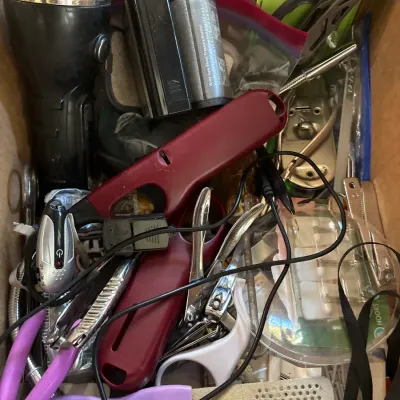 Cardboard box filled with assorted household items, including a black flashlight, utility lighters, nail clippers, tools, and small...