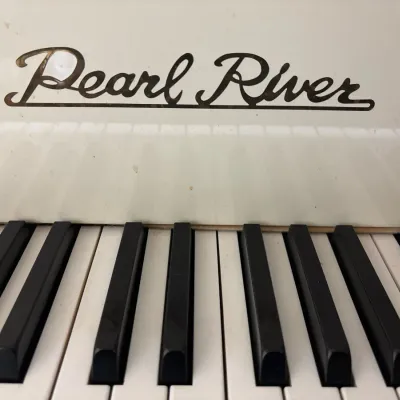 Close-up of a Pearl River white piano, highlighting the gold cursive brand logo above the keyboard.