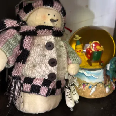 Plush snowman figure dressed in a knit sweater, scarf, and hat holding ice skates, displayed next to a tropical-themed snow globe.