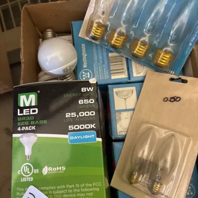 Box filled with assorted lightbulbs, including a 4-pack of M LED BR30 8W daylight bulbs, packaged decorative candelabra bulbs, and...