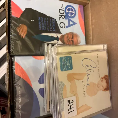 Cardboard box containing a collection of CDs and DVDs, including Celine Dion's 'Falling Into You' CD and Dr. Steven Gundry...
