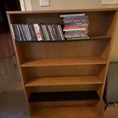 Tall wood laminate media storage rack holding a small collection of CDs and VHS tapes on the top shelf.