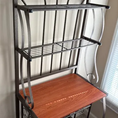 Contemporary metal baker's rack featuring decorative scrollwork, two wire shelves, a lower wine glass rack, and a wood-tone prep...