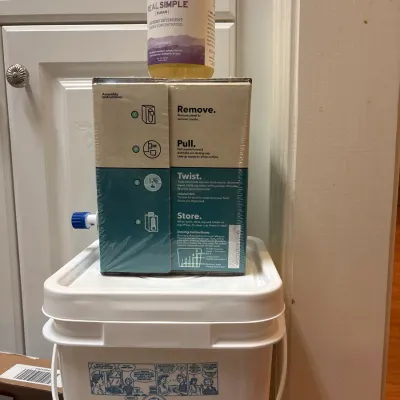 Stack of laundry supplies, including a white plastic bucket with lid, a boxed liquid detergent with dispenser nozzle, and a bottle of...