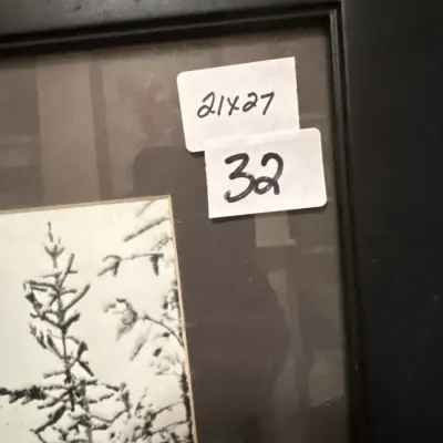 Framed black-and-white landscape artwork featuring snow-covered evergreen trees, measuring 21 by 27 inches.