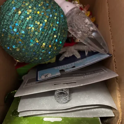 Cardboard box filled with assorted Christmas decorations, including a large green sequined sphere ornament and packaged Holiday Time mini...
