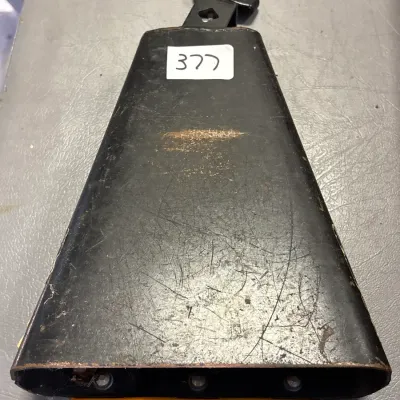 Black metal percussion cowbell with a mounting bracket and a yellow plastic edge guard, showing visible surface scuffs and wear.