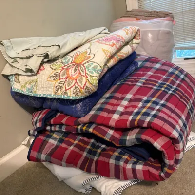 Stack of folded bedding linens, including a red plaid comforter, a embroidered floral quilt, and various blankets.