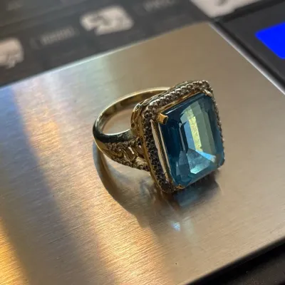 Gold-tone cocktail ring featuring a large emerald-cut blue topaz center stone surrounded by a halo of small clear accent stones, resting...