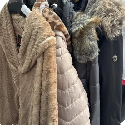 Selection of women's winter coats and jackets hanging on a rack, featuring faux fur wraps, quilted puffer coats, and faux...