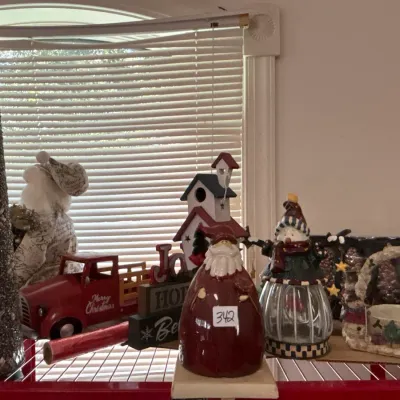Collection of holiday table decor including a red ceramic Santa figurine, a snowman lantern, a red toy truck, a sparkly cone tree, and a...