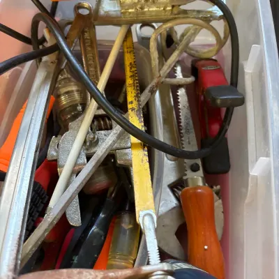 Plastic storage container filled with assorted hand tools and power tools, including an orange electric jigsaw, hacksaws, wrenches, and...