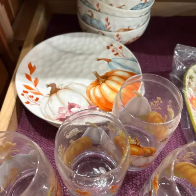 Autumn-themed tableware set including pumpkin motif ceramic plates, stacked bowls, and matching drinking glasses.