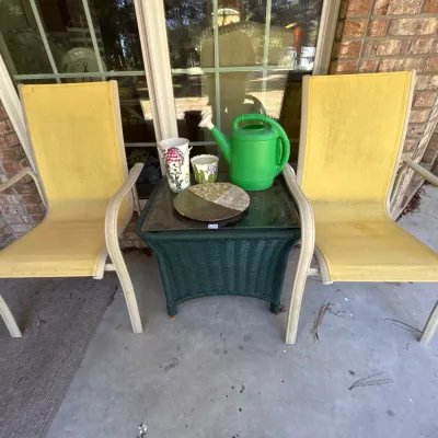 Pair of yellow sling patio chairs alongside a green woven resin side table topped with a plastic watering can, floral vases, and...