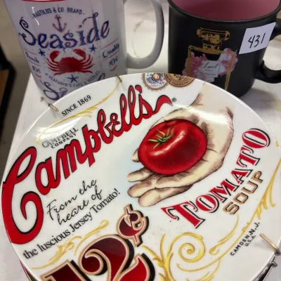 Vintage Campbell's Tomato Soup collectible ceramic plate displayed on a wall hanger alongside two coffee mugs, including a Seaside...