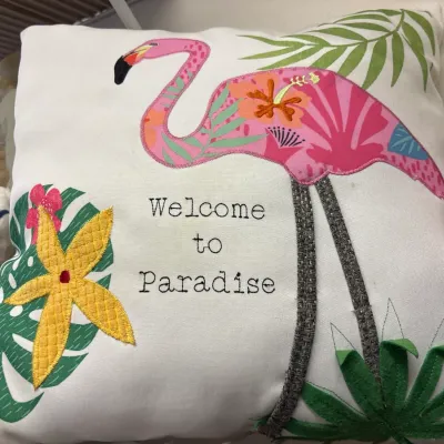 Square white throw pillow featuring embroidered flamingo, palm leaves, tropical flower appliqué, and the text "Welcome to...