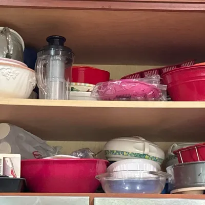 Assorted kitchenware and bakeware stored on wooden pantry shelves, including mixing bowls, a manual food chopper, and a boxed electric...