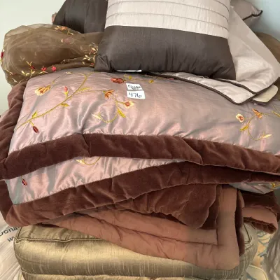 Stack of bedding and decorative pillows featuring a satin floral comforter with brown velvet trim, a pleated accent pillow, and...
