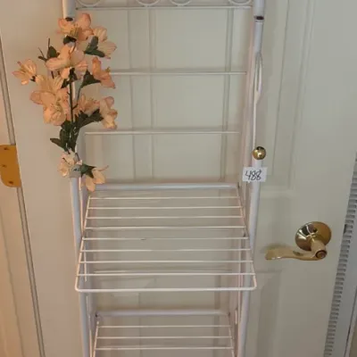 Tall white painted metal wire rack featuring tiered shelving scrollwork top detail and faux flower decorations.