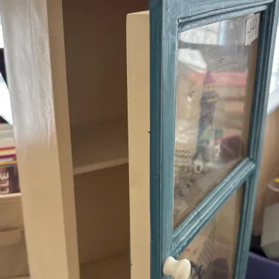 Small wooden wall cabinet featuring blue-painted trim and glass door panels displaying nautical decorative motifs.