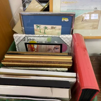 Collection of assorted picture frames and artwork stored in a green plastic bin, including silver-tone and wooden frames with...