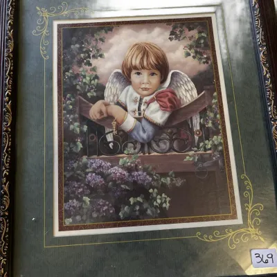 Framed artwork depicting a young cherubic angel leaning over a balcony surrounded by flowers, presented in an ornate dark wood frame with...