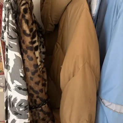 Collection of winter coats and jackets hanging on a rack, including a tan hooded puffer jacket, leopard print faux fur coat, and a light...