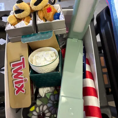 Box lot of assorted small household items, featuring plush toys in boxes, a Twix candy tin, a stained glass flower ornament, a folding...