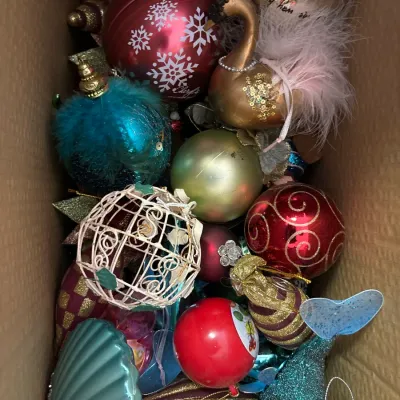 Cardboard box filled with an assortment of vintage and modern Christmas tree ornaments, including a feathered swan, glittery seashells,...