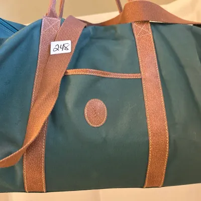 Vintage dark green canvas duffel bag featuring brown webbed and leather trim, top handles, an adjustable shoulder strap, and an oval...